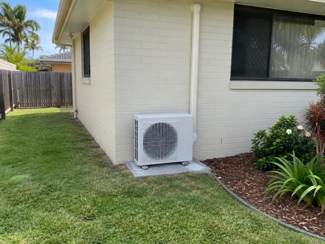 Outdoor air conditioner unit on a concrete pad beside a cream brick house with pipe cover in Tugun