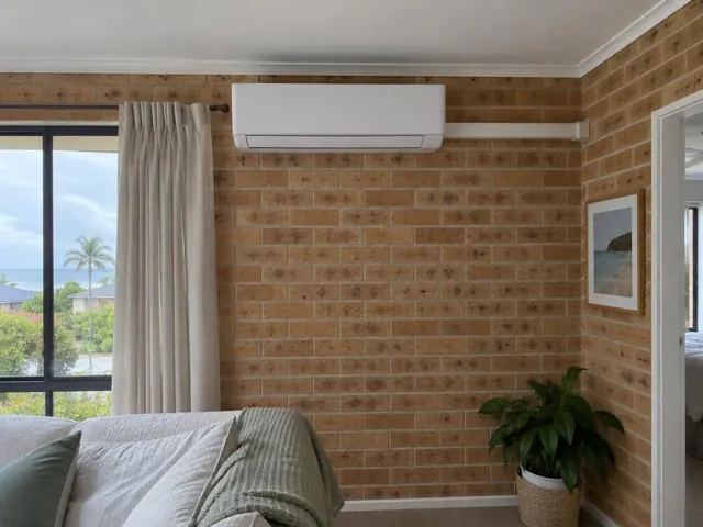 Wall-mounted split system on a brick feature wall in a living room with curtains and window in Currumbin Waters